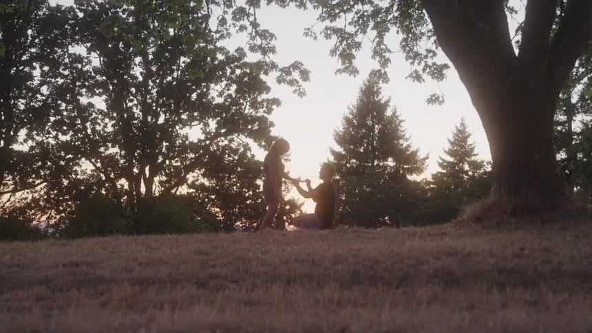 Man kneels proposing to woman in park at sunset