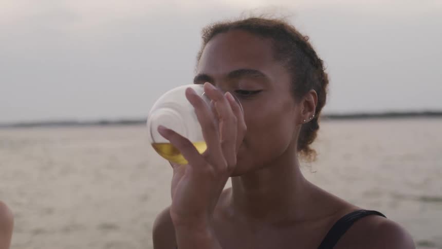 A woman with dark hair pulled back in a ponytail sips from a glass of white wine while sitting on a sandy beach