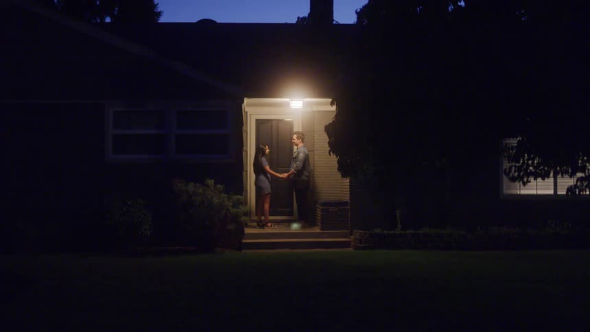 A woman and a man are walking up the steps to a house at dusk they stop at the door hold hands and share a brief intimate moment before the man walks away
