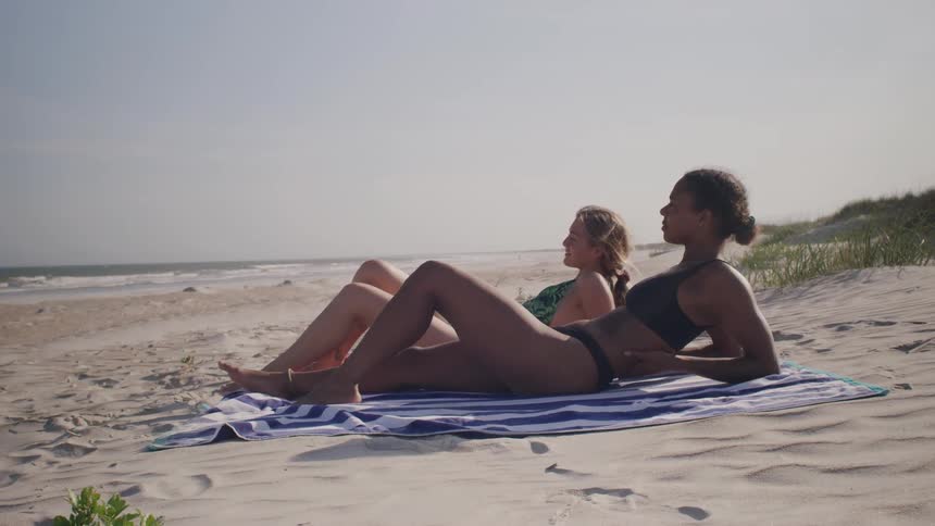 A woman in a dark bikini and a woman in a green and black one piece bathing suit lie on a striped towel on a beach