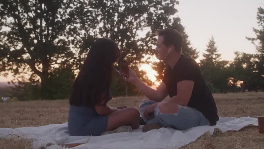 A man and a woman sit on a blanket on a grassy hill sharing earbuds and smiling at each other as the sun sets behind them