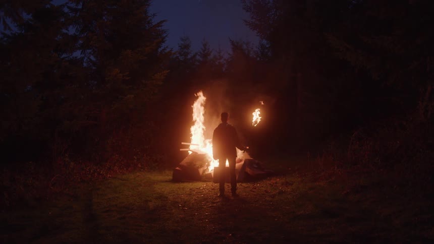 A person holding a torch stands in front of a large bonfire surrounded by forest at night