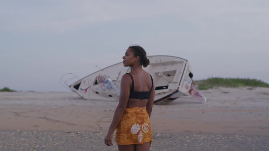 A young girl wearing a mini skirt walks on the beach with a boat on the sand ahead as the wind blows her skirt at midday