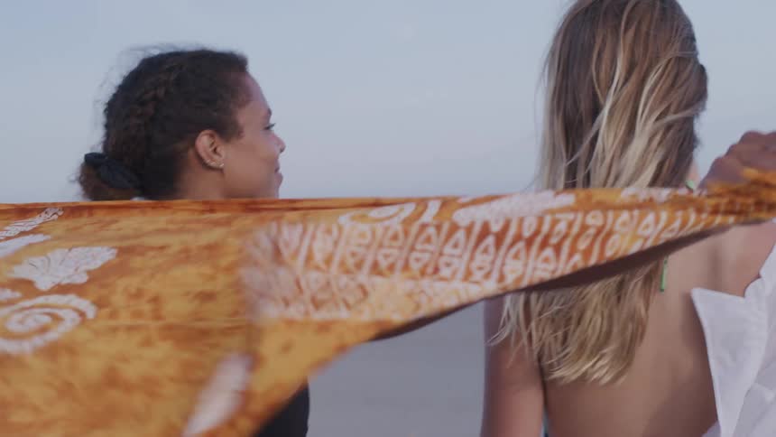 A woman with dark hair pulled back and a woman with long blonde hair walk on a beach facing the ocean with the woman on the left smiling and holding a patterned orange towel