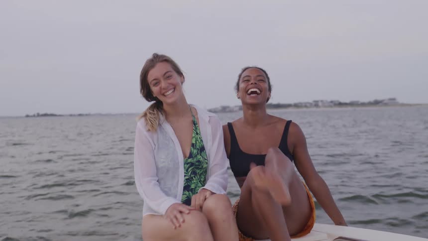 The video shows two young women on a boat sitting on the deck with a large body of water and land in the background they are both smiling looking at the camera and raising their arms to make a peace sign appearing to be enjoying their time together