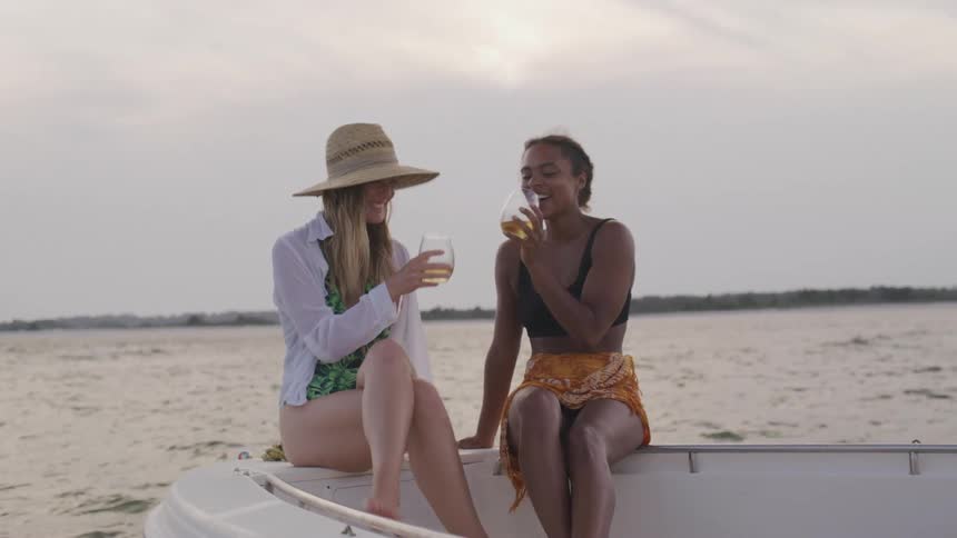 Two women smile and toast drinks together on a boat at sunset