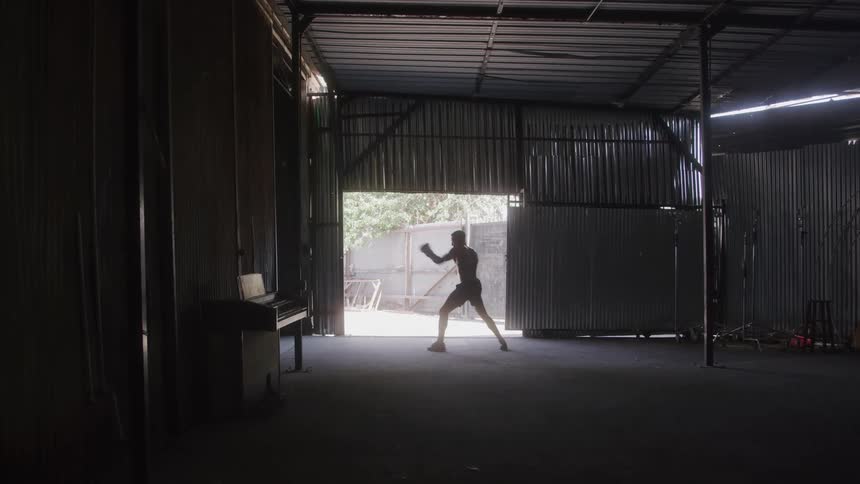 A man shadowboxes in a dimly lit warehouse with sunlight streaming through the open door behind him