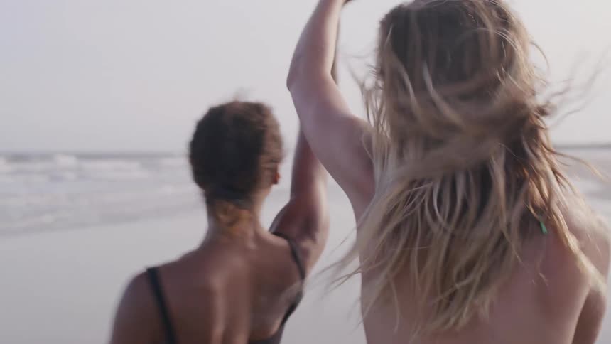 Two women one with her hair up and the other with long blonde hair are walking and laughing together on a beach with their backs mostly to the camera