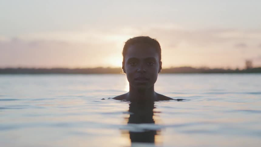 The video shows a person in a body of water with their shoulders and head above the surface the persons face is in shadow as they look directly at the camera