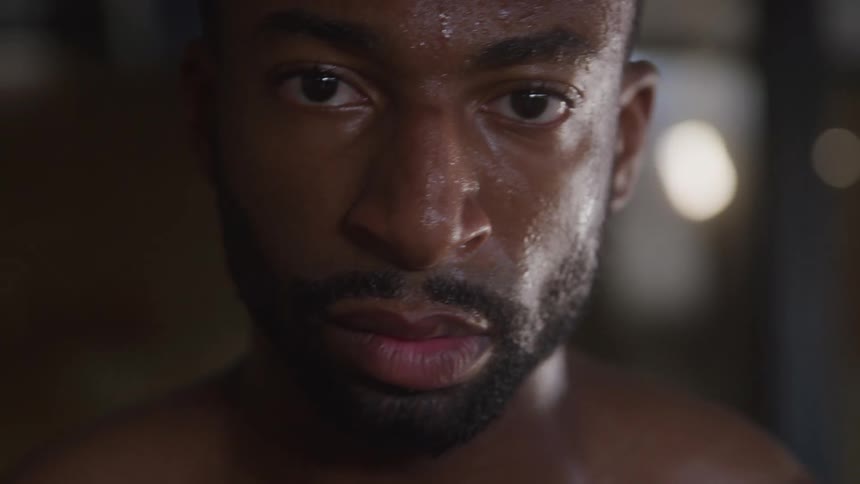 The video shows a very closeup shot of a mans face who appears to be sweating heavily he looks directly into the camera with a determined expression and the low lighting suggests he is in a gym or engaged in an intense workout