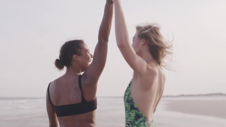 Two women in swimsuits walk along the beach one holding a flowing orange cloth