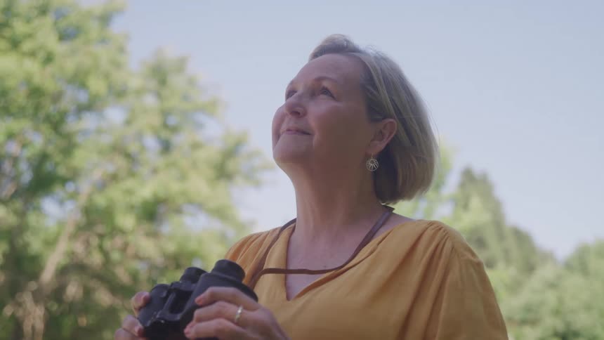 Woman in a yellow blouse holds binoculars and looks up outdoors