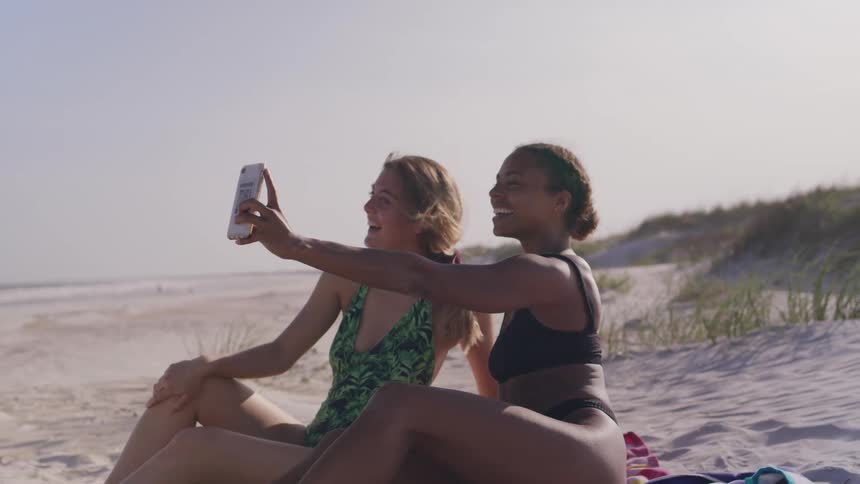 Two women sitting on the sand take a smiling selfie at the beach