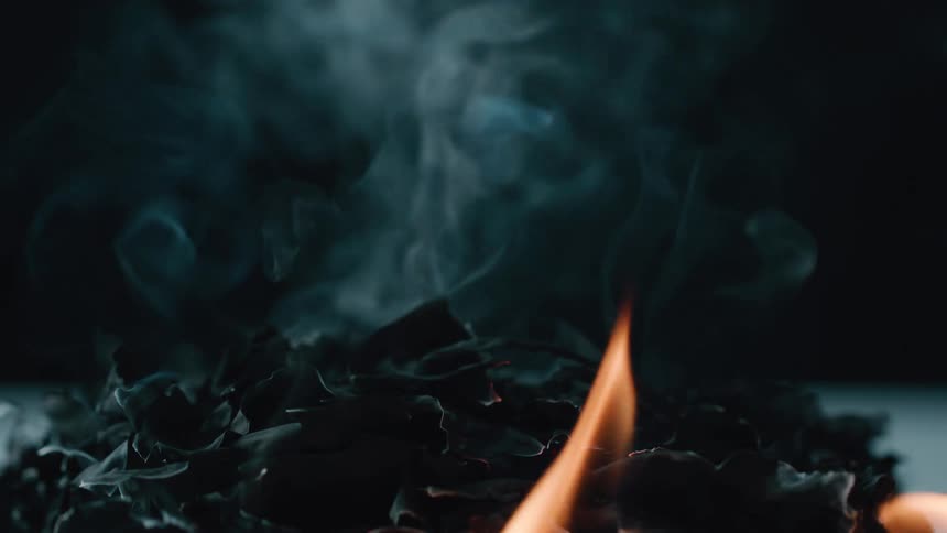 Dark smoke rises from a pile of burnt or charred material in low light