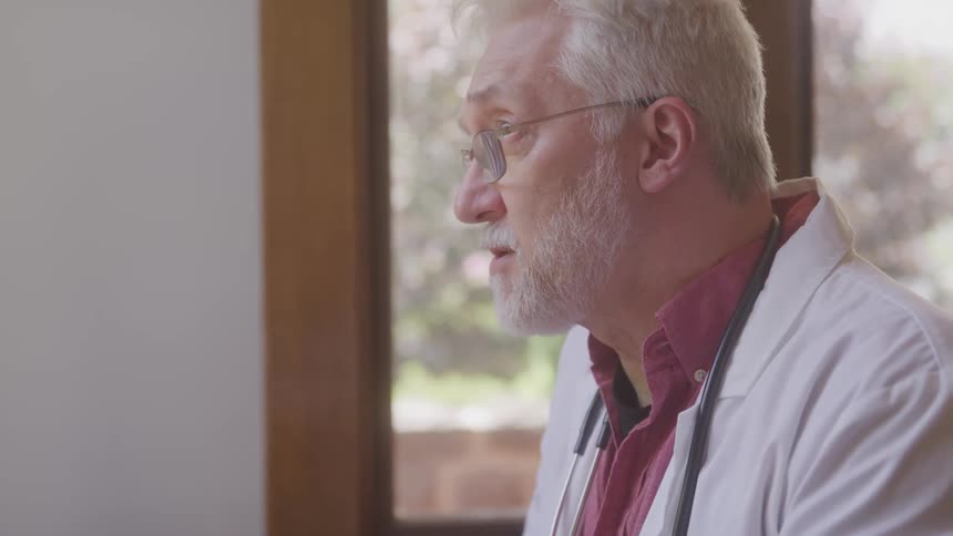 A doctor with gray hair and a beard is speaking animatedly he is wearing a white lab coat over a red shirt and has a stethoscope around his neck