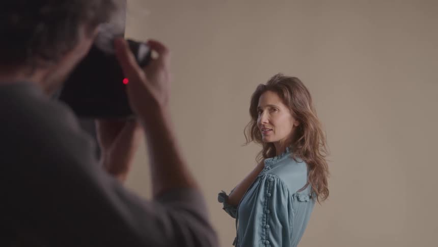The video shows a photographer taking photos of a woman with long brown hair the woman is wearing a denim shirt and looks directly at the camera with her arms crossed