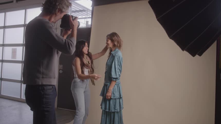 A person is standing in a studio with a photographer and a second person the photographer is holding a camera and the second person is adjusting the models hair