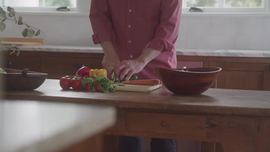 A person wearing a pink long sleeved shirt stands in a kitchen and chops vegetables on a cutting board with a wooden bowl and a pile of vegetables on the table in front of him