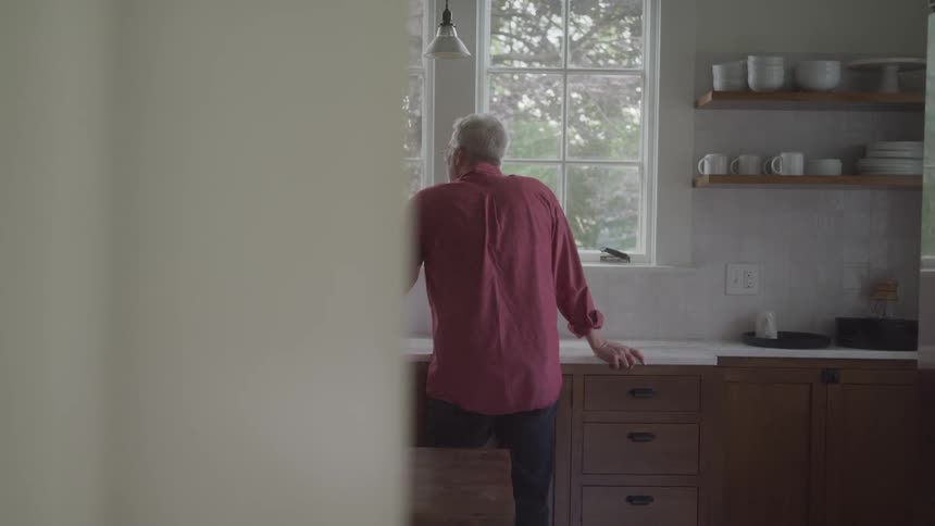 An older man stands in a kitchen facing a window