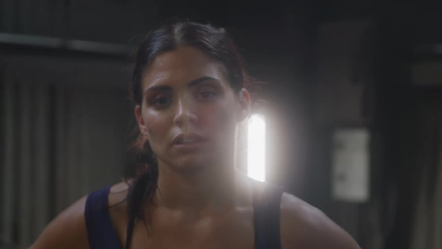 A mediumclose up of a woman with a serious expression looking directly at the camera in a dimly lit space likely a gym