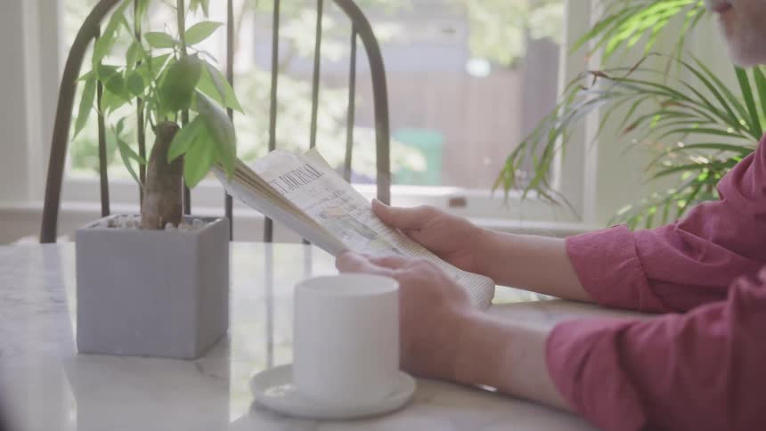 A person reads a newspaper at a table near a window with houseplants