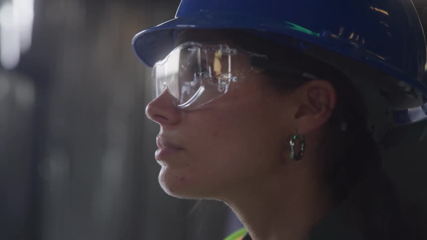 The video shows a closeup of a womans face wearing a blue hard hat and safety glasses looking intently at something offscreen the woman is a construction worker or engineer as suggested by her attire