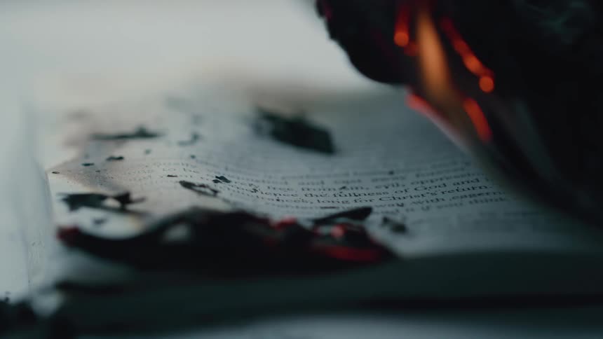The charred edges of a burning book curl upward as faint text remains visible amid glowing embers