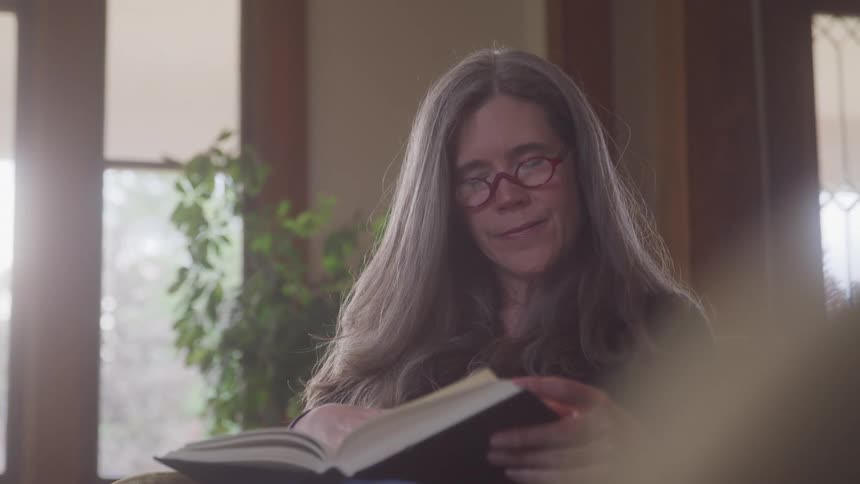 A woman with long gray hair and red glasses reads a book in a softly lit living room