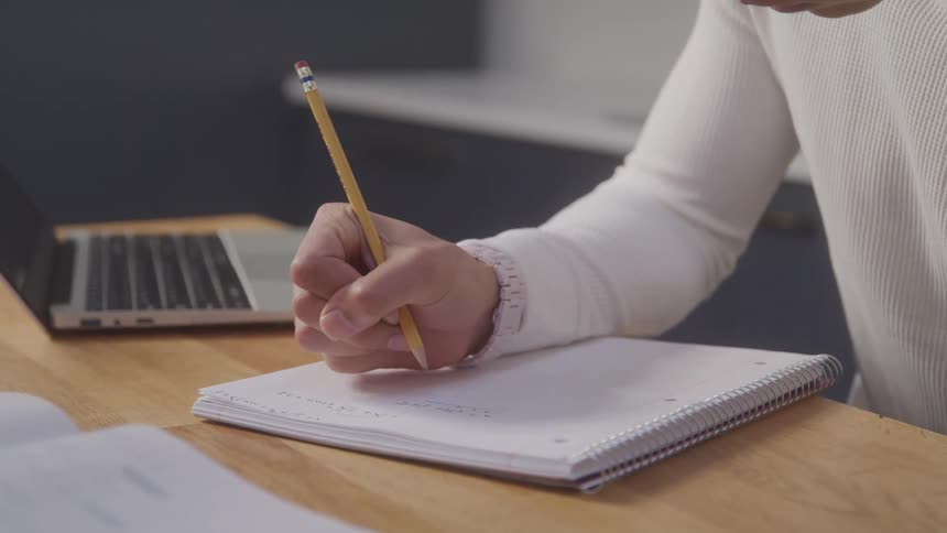 A man is writing in a notebook with a pencil and theres a laptop on the table that hes studying