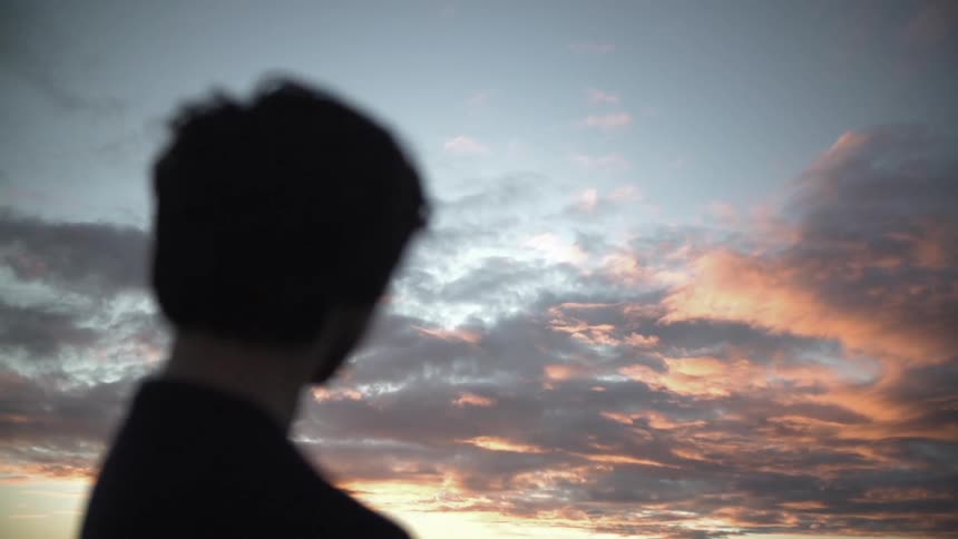 A person whose image is blurred is looking towards a beautiful sunset with the reddish light shining on the clouds