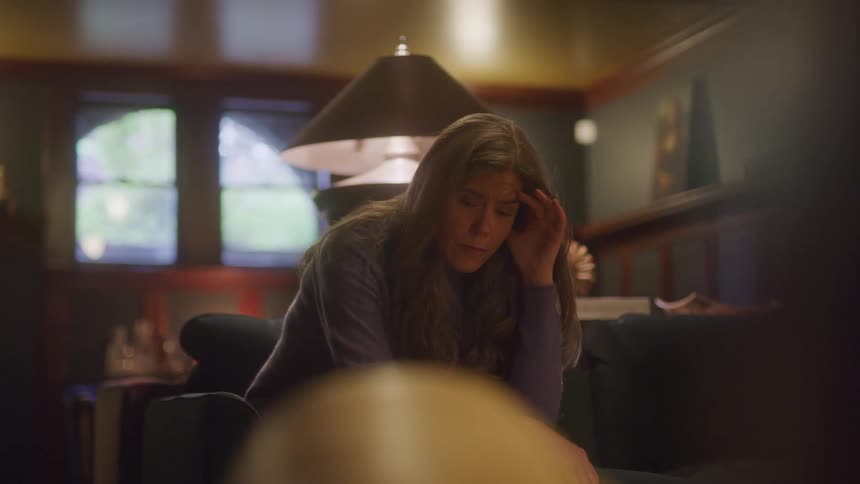 A woman with long dark hair is sitting on a couch in a dimly lit room with a solemn expression on her face