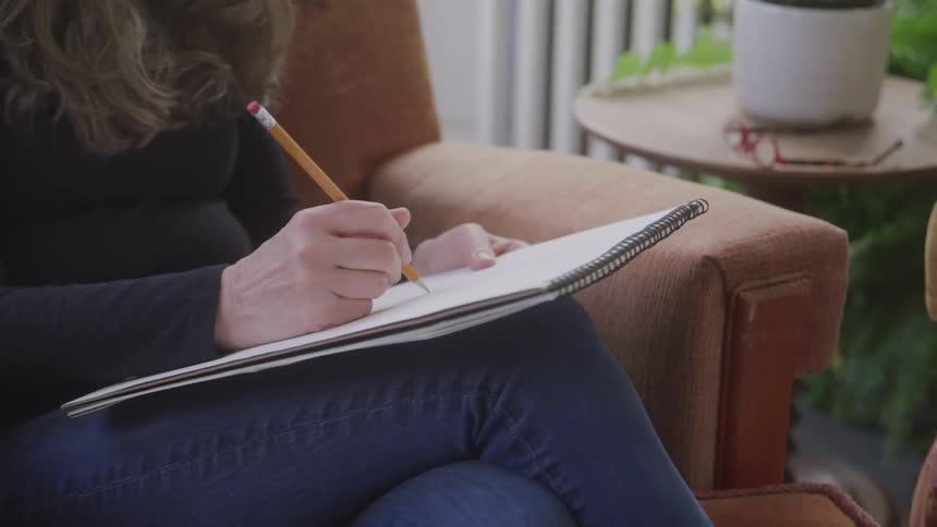 The video shows a closeup of a persons hands holding a pencil and an open spiral notebook they are sitting in a comfortable armchair and their hands are actively writing or sketching in the book