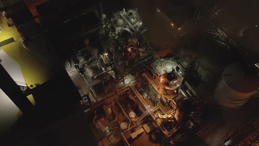 Thick industrial smoke billows from a large complex facility lit by bright floodlights at night