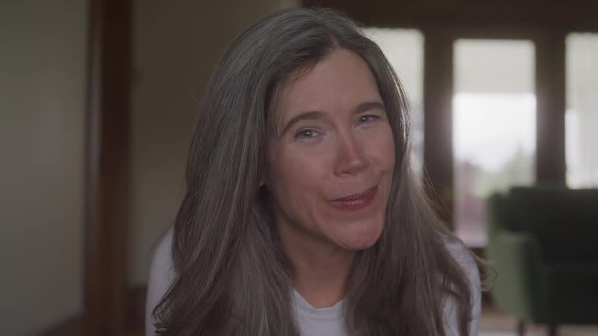 A woman with long graying hair and a faint smile on her face looks directly at the camera the background is a dimly lit indoor space with a window and a sofa visible behind her