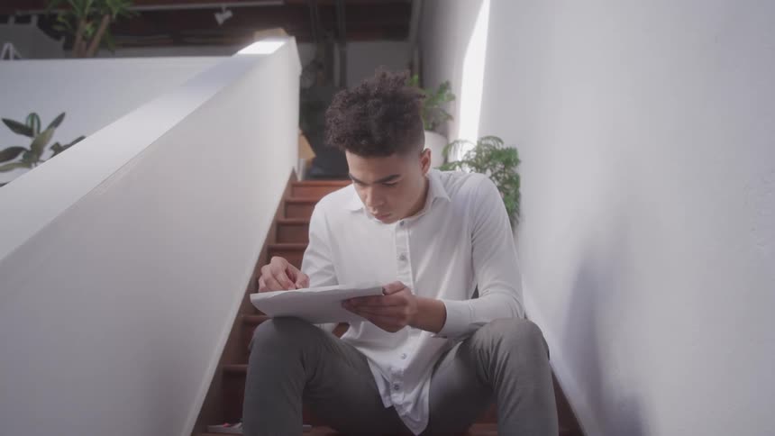A young man in a white shirt sits on stairs focused on reading and writing