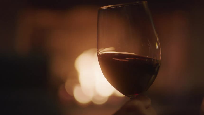 A hand holds a glass of red wine glows softly before a cozy fireplace
