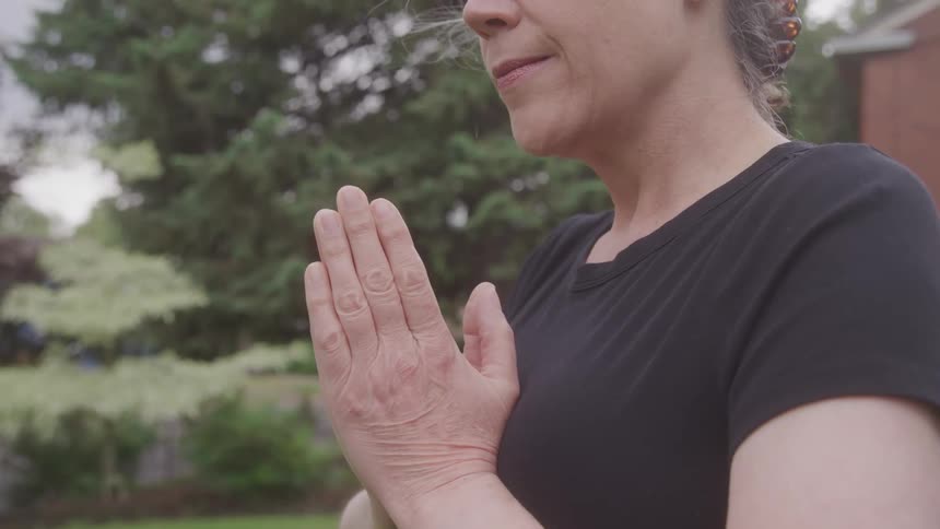 A peacefullooking woman with a hint of a smile is standing outdoors holding her hands together in a prayer pose with a large green tree in the background