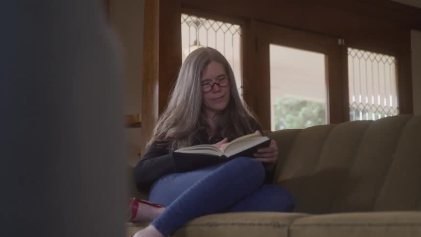 The video shows a woman with glasses and long hair sitting on a couch engrossed in a book she is wearing a dark top jeans and red shoes and is sitting comfortably in a welllit living room