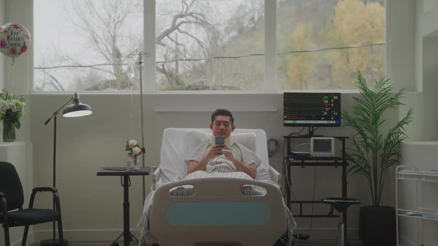 A male patient is lying in a hospital bed looking at his phone with a small smile with a get well soon balloon visible behind him
