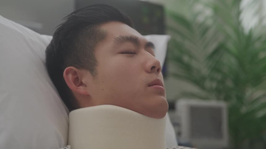 A closeup shot of a young man lying peacefully with his eyes closed in a hospital bed wearing a neck brace and appearing to be asleep