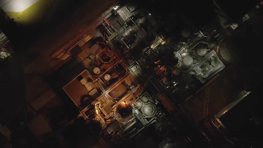 An aerial shot shows an industrial facility at night with various buildings and interconnected pipes the scene is illuminated by scattered lights that cast a glow on the structures below