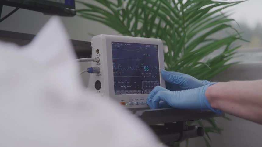 A medical professional wearing blue gloves adjusts a medical monitor that displays vital signs while a plant sits on a table nearby