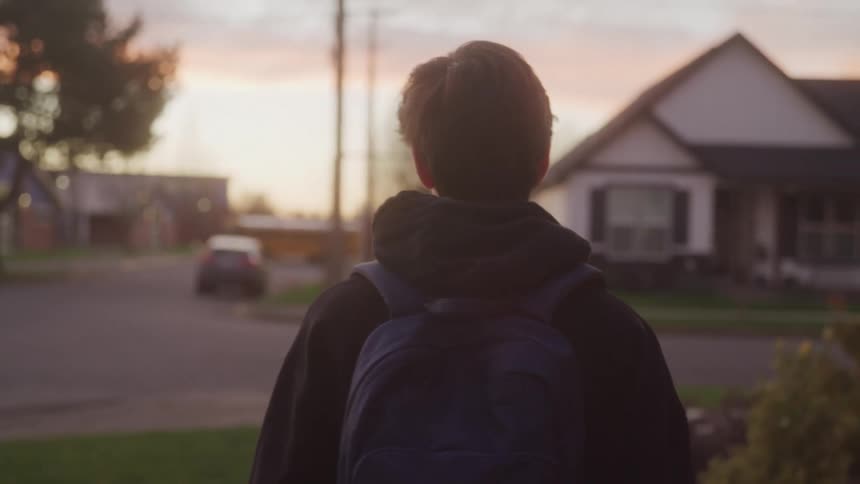 A cinematic overtheshoulder shot of a person with a backpack walking down a suburban street at dusk or dawn