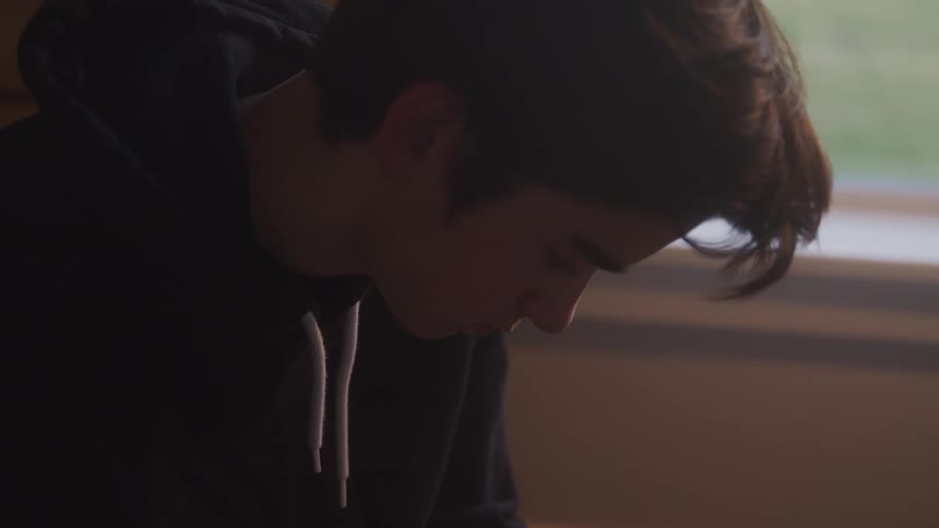 A young man in a dark hoodie is seen in a dimly lit room with his head bowed down in a pensive or somber manner