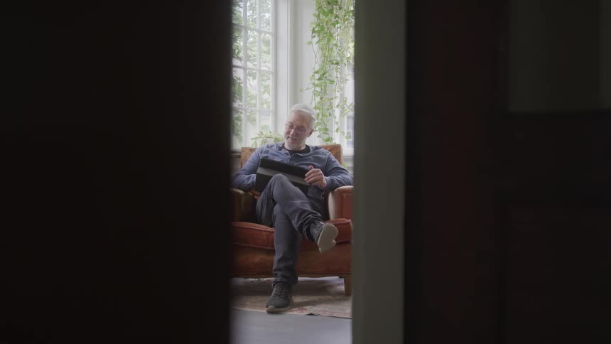 The video shows an older man with gray hair sitting in a comfortable armchair seen through a partially open doorway he is holding a book or tablet and appears to be smiling and relaxed enjoying the quiet moment