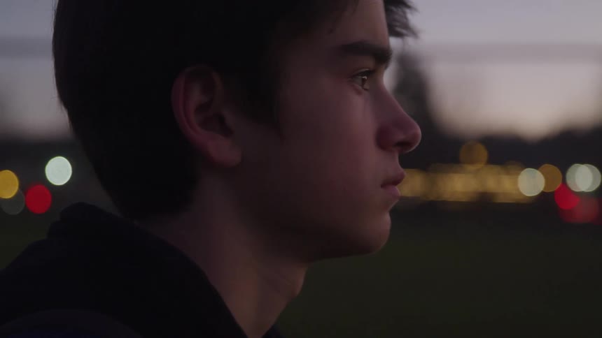 A teenage boy with short brown hair stands outdoors at dusk looking down and then up with a serious expression