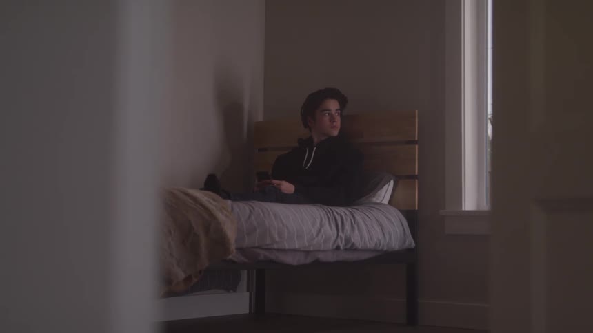 A teenager wearing a dark hoodie and headphones sits up in bed looking out a window
