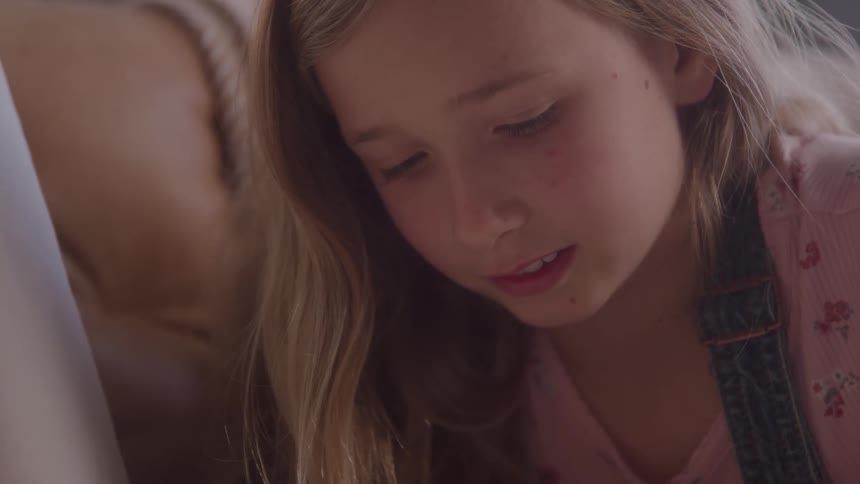 The video shows a closeup of a young girl with long blonde hair looking down with a focused expression she is wearing a pink shirt and blue overalls with a floral pattern suggesting she is engaged in an activity that requires her concentration