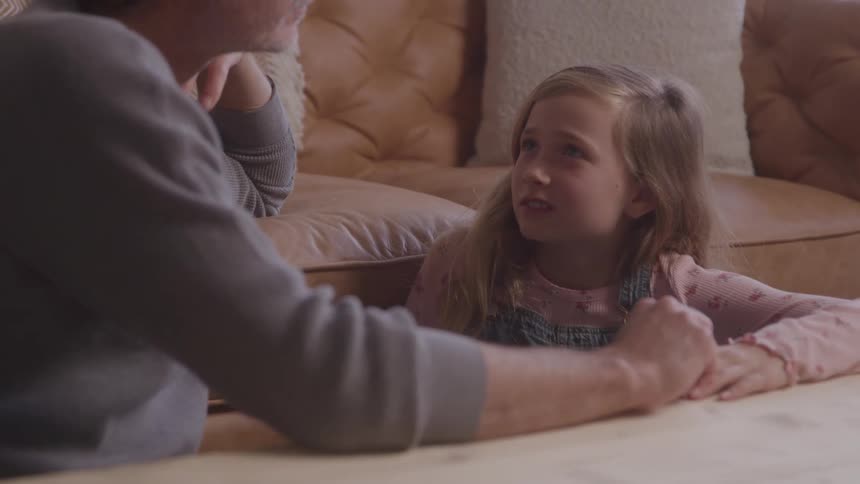 A man is kneeling on the floor holding the hands of a young girl with long blonde hair they are talking to each other with warm and caring expressions