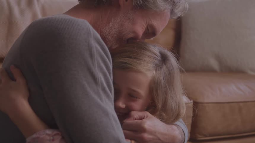 A man hugs a smiling young girl tightly in a tender moment on a couch